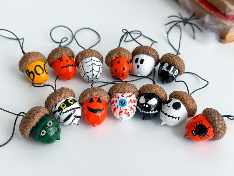 K&ouml;nnte beinhalten: Eine Girlande aus 13 Halloween-f&ouml;rmigen Eicheln, die mit verschiedenen gruseligen Designs bemalt sind, darunter ein Geist, eine Spinne, eine Mumie, ein K&uuml;rbis und ein Sch&auml;del. Jede Eichel ist an einem schwarzen Draht befestigt und hat oben eine kleine Schlaufe zum Aufh&auml;ngen.