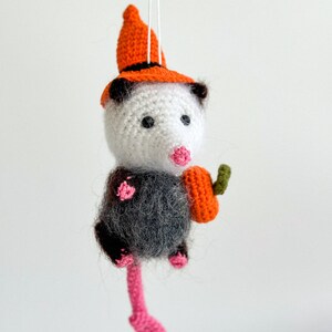 May include: A crocheted opossum wearing an orange and black witch hat and holding a small orange pumpkin. The opossum has a pink nose and black eyes.