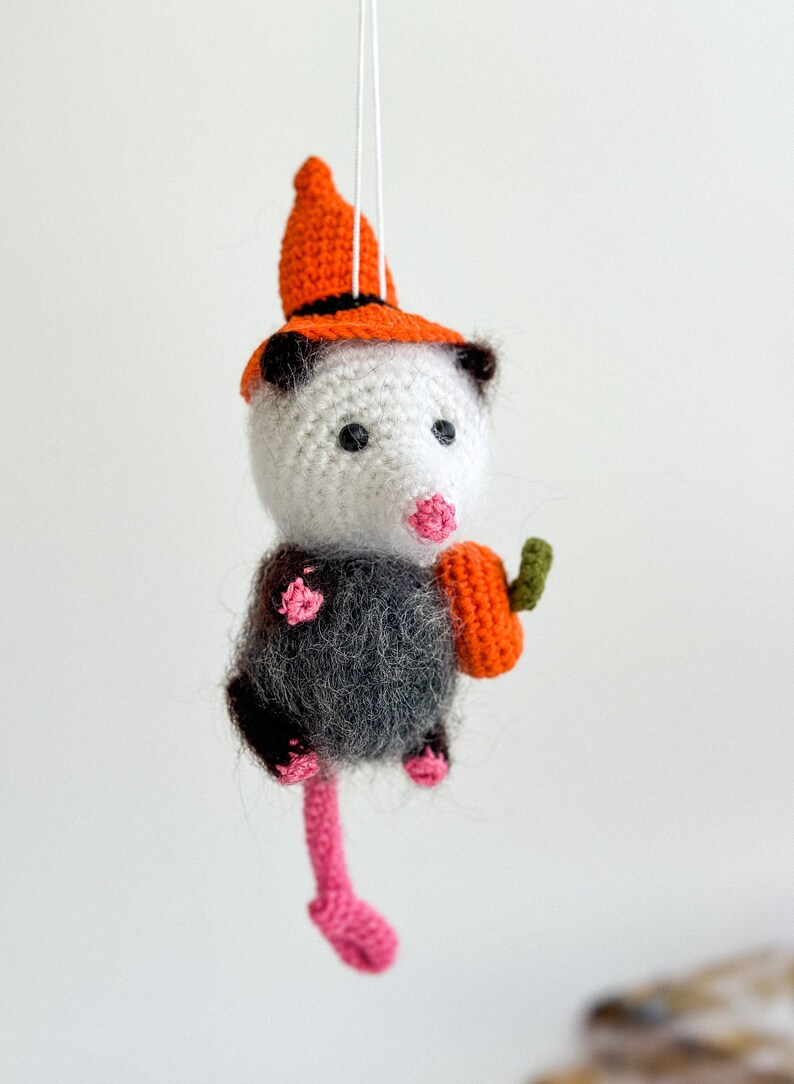 May include: A crocheted opossum wearing an orange and black witch hat and holding a small orange pumpkin. The opossum has a pink nose and black eyes.
