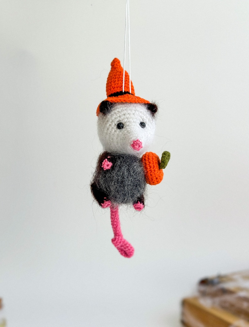 May include: A crocheted opossum wearing an orange witch hat and holding a small orange pumpkin. The opossum has black and white fur and a pink nose.