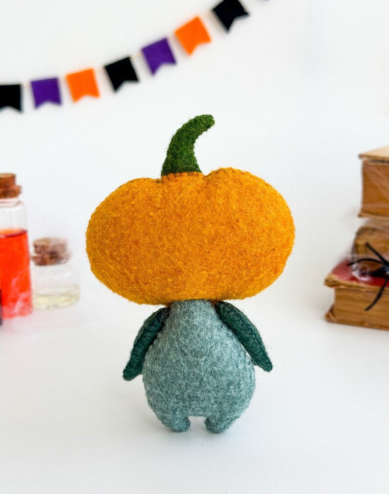 May include: A felt pumpkin character with a green stem and a teal body. The pumpkin is orange and has a fuzzy texture.