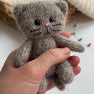 K&ouml;nnte beinhalten: Eine kleine, graue, gestrickte Katzendoll mit schwarzen Augen, Schnurrhaaren und einer rosa Nase. Das handgefertigte Spielzeug hat ein schlichtes Design und wird in einer Hand gehalten. Die Ohren der Katze sind abgerundet und der K&ouml;rper ist weich und pl&uuml;schig.