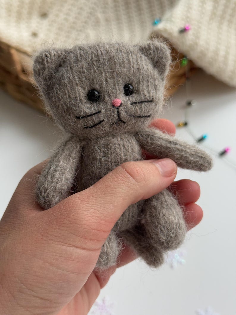 K&ouml;nnte beinhalten: Eine kleine, graue, gestrickte Katzendoll mit schwarzen Augen, Schnurrhaaren und einer rosa Nase. Das handgefertigte Spielzeug hat ein schlichtes Design und wird in einer Hand gehalten. Die Ohren der Katze sind abgerundet und der K&ouml;rper ist weich und pl&uuml;schig.