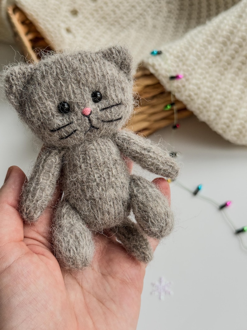 K&ouml;nnte beinhalten: Eine kleine, graue, gestrickte Katzendoll mit schwarzen Augen, Schnurrhaaren und einer rosa Nase. Das handgefertigte Spielzeug wird in einer Hand gehalten und zeigt seine weiche Textur und detaillierte Handwerkskunst. Die Ohren der Katze sind aufrecht und ihr K&ouml;rper ist kompakt.
