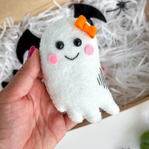 May include: A white felt ghost with black eyes, a pink blush, and a black stitched smile. The ghost has an orange felt bow and is held in a hand against a white background.