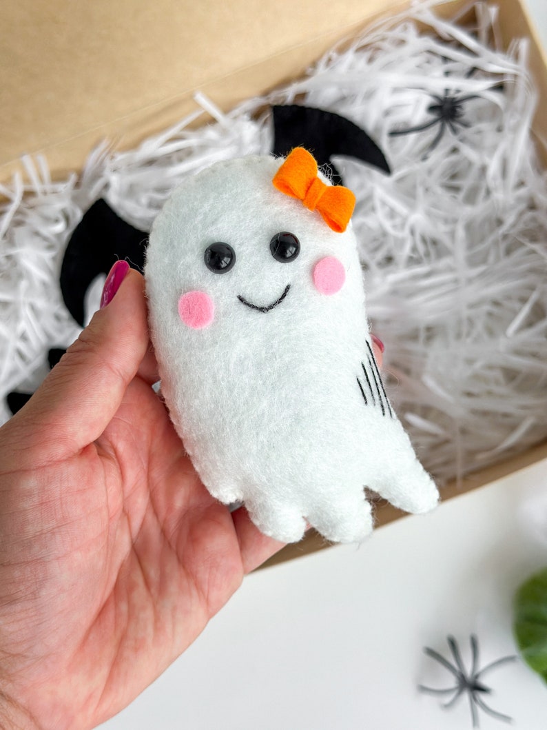 May include: A white felt ghost with black eyes, a pink blush, and a black stitched smile. The ghost has an orange felt bow and is held in a hand against a white background.