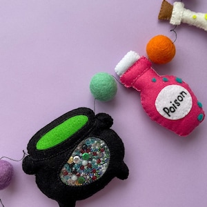 K&ouml;nnte beinhalten: Eine Halloween-Girlande mit Filzdekorationen, darunter ein Kessel gef&uuml;llt mit bunten Pailletten, eine rosa Zaubertrankflasche mit dem Wort "Gift" darauf und gr&uuml;ne, orange und violette Filzkugeln.