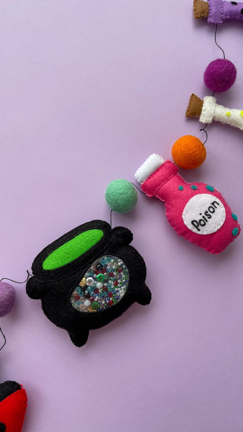 K&ouml;nnte beinhalten: Eine Halloween-Girlande mit Filzdekorationen, darunter ein Kessel gef&uuml;llt mit bunten Pailletten, eine rosa Zaubertrankflasche mit dem Wort "Gift" darauf und gr&uuml;ne, orange und violette Filzkugeln.