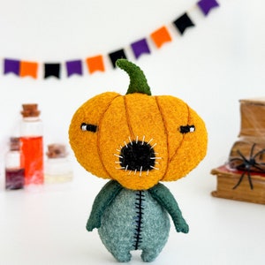 May include: A felt pumpkin character with a stitched mouth and green felt body. The pumpkin is orange and has a green stem. The character is standing on a white surface.