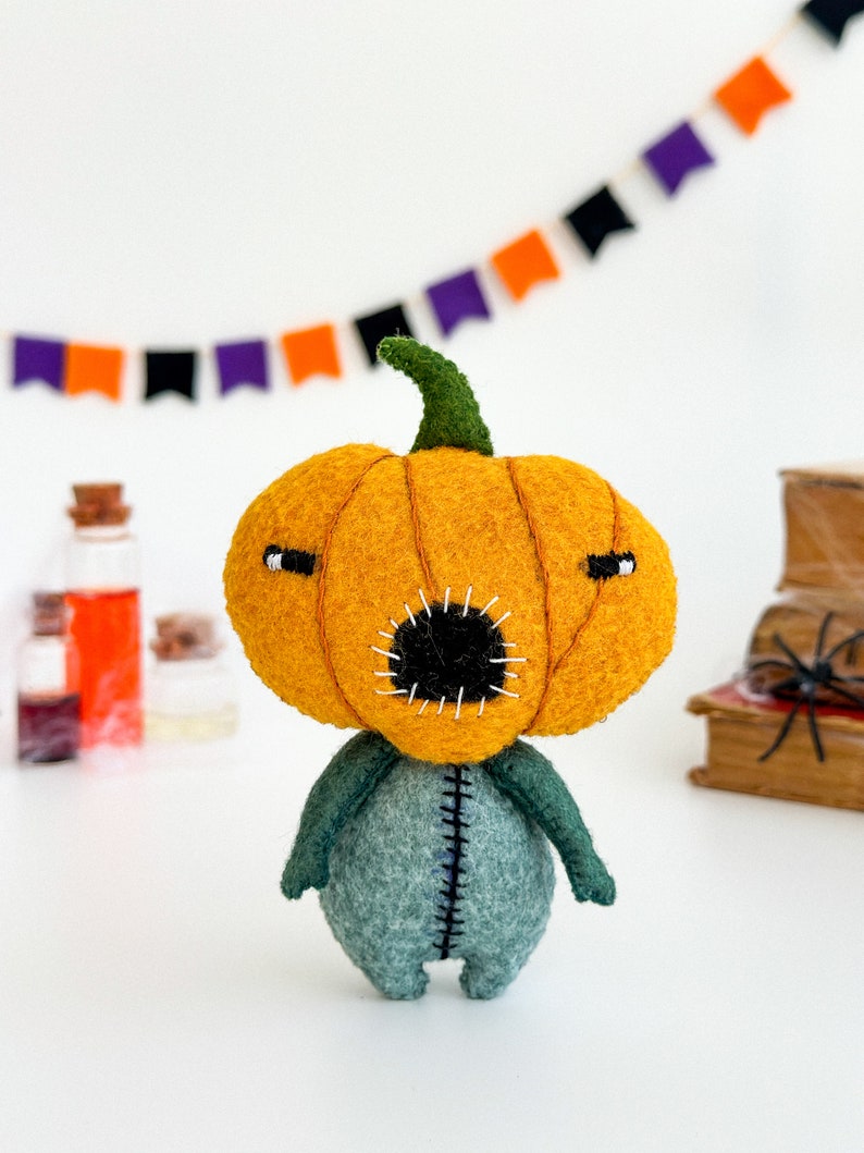 May include: A felt pumpkin character with a stitched mouth and green felt body. The pumpkin is orange and has a green stem. The character is standing on a white surface.