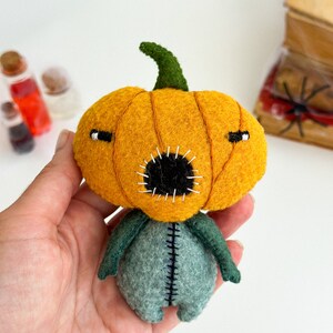 May include: A felt pumpkin doll with a stitched mouth and eyes. The doll is wearing a green felt body with stitched seams.
