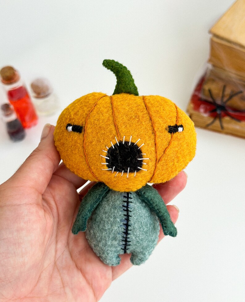May include: A felt pumpkin doll with a stitched mouth and eyes. The doll is wearing a green felt body with stitched seams.