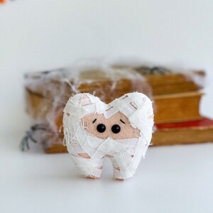 May include: A plush tooth wrapped in white fabric bandages, with black button eyes, resembling a mummy. The tooth is set against a blurred background with books and Halloween decorations, suggesting a spooky theme.
