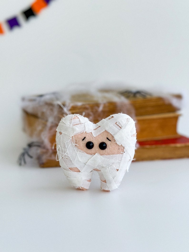 May include: A plush tooth wrapped in white fabric bandages, with black button eyes, resembling a mummy. The tooth is set against a blurred background with books and Halloween decorations, suggesting a spooky theme.