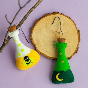 Puede incluir: Dos botellas de poci&oacute;n de fieltro, una amarilla con una calavera y huesos cruzados y la otra verde con una luna creciente y estrellas, colgando de una rama y una rodaja de madera.
