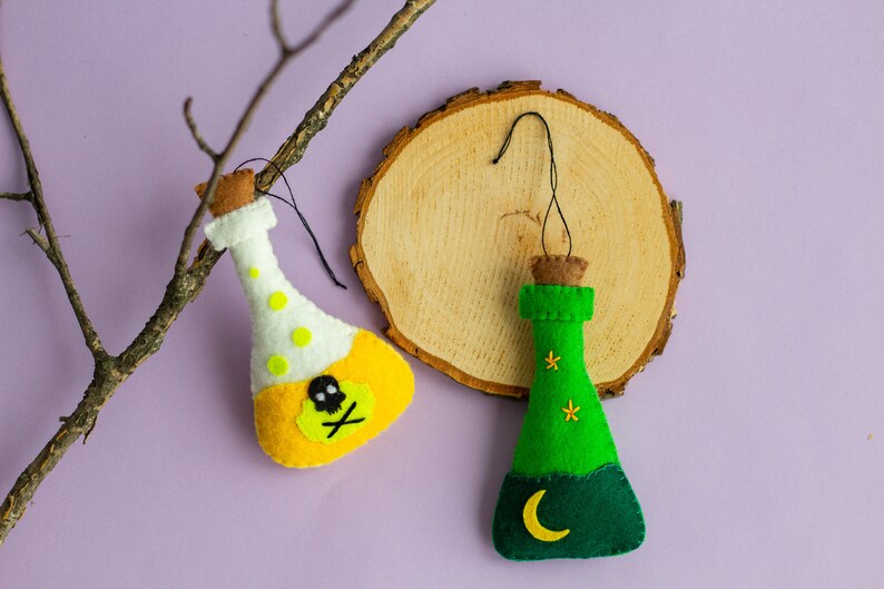Puede incluir: Dos botellas de poci&oacute;n de fieltro, una amarilla con una calavera y huesos cruzados y la otra verde con una luna creciente y estrellas, colgando de una rama y una rodaja de madera.