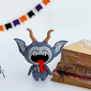 May include: A gray felt monster with horns and a red tongue sticking out. The monster is smiling and has a mischievous expression. The monster is sitting on a stack of old books with a spider web on them.