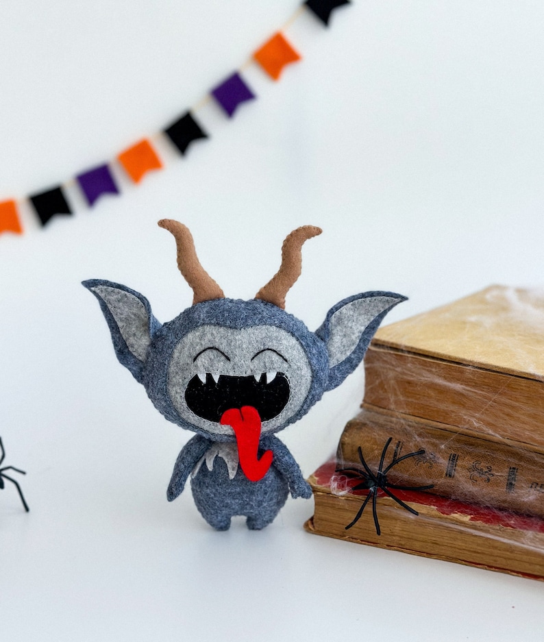 May include: A gray felt monster with horns and a red tongue sticking out. The monster is smiling and has a mischievous expression. The monster is sitting on a stack of old books with a spider web on them.