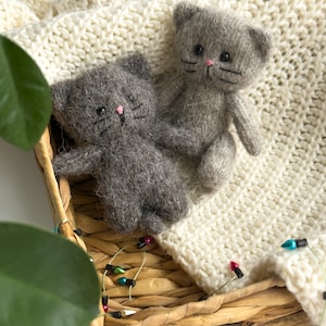 K&ouml;nnte beinhalten: Zwei graue Katzenpl&uuml;schtiere mit rosa Nasen und schwarzen Schnurrhaaren liegen in einem geflochtenen Korb, der mit einer cremefarbenen Strickdecke ausgekleidet ist. Eine Lichterkette mit bunten Lichtern schm&uuml;ckt den Rand des Korbes und verleiht der Szene eine festliche Note.