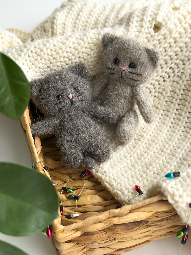 K&ouml;nnte beinhalten: Zwei graue Katzenpl&uuml;schtiere mit rosa Nasen und schwarzen Schnurrhaaren liegen in einem geflochtenen Korb, der mit einer cremefarbenen Strickdecke ausgekleidet ist. Eine Lichterkette mit bunten Lichtern schm&uuml;ckt den Rand des Korbes und verleiht der Szene eine festliche Note.