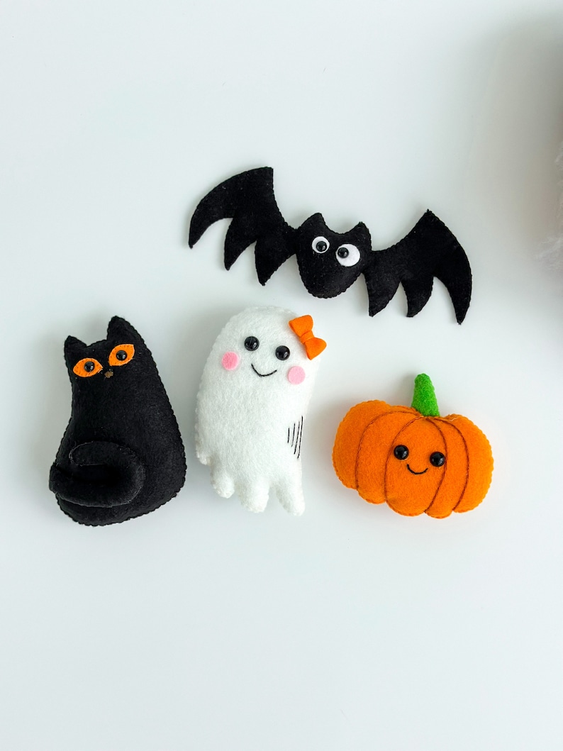 May include: A set of four felt Halloween decorations: a black cat, a white ghost with a pink bow, an orange pumpkin, and a black bat.