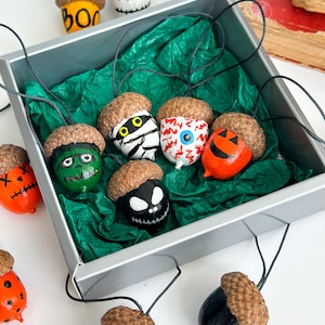 K&ouml;nnte beinhalten: Eine Sammlung von neun Eichel-Ornamenten, die mit verschiedenen Halloween-Charakteren bemalt sind, darunter eine Mumie, ein Geist, ein K&uuml;rbis und ein Zombie. Die Eicheln sind mit schwarzem Garn zusammengef&uuml;gt und in einer silbernen Schachtel mit gr&uuml;nem Seidenpapier ausgelegt.