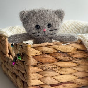 K&ouml;nnte beinhalten: Ein handgefertigtes graues Katzenpl&uuml;schtier mit schwarzen Augen und rosa Nase blickt aus einem geflochtenen Korb. Der Korb ist mit einer wei&szlig;en Strickdecke ausgekleidet. Dekorative Lichterketten mit bunten Gl&uuml;hbirnen sind um den Korb drapiert.