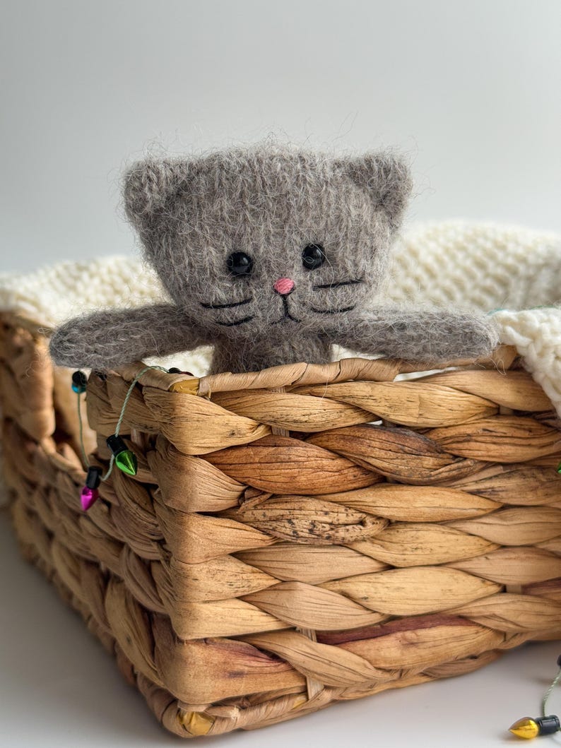 K&ouml;nnte beinhalten: Ein handgefertigtes graues Katzenpl&uuml;schtier mit schwarzen Augen und rosa Nase blickt aus einem geflochtenen Korb. Der Korb ist mit einer wei&szlig;en Strickdecke ausgekleidet. Dekorative Lichterketten mit bunten Gl&uuml;hbirnen sind um den Korb drapiert.