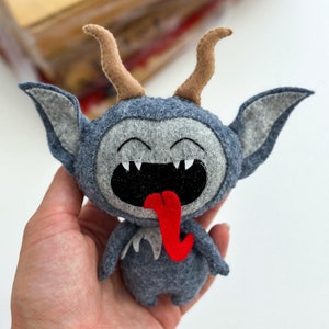 May include: A gray felt plush toy with a red tongue sticking out. The toy has brown horns and a wide, toothy grin.