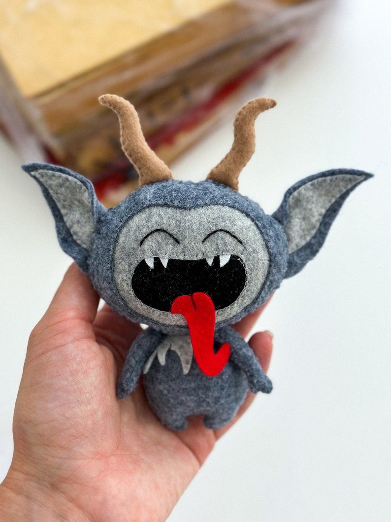 May include: A gray felt plush toy with a red tongue sticking out. The toy has brown horns and a wide, toothy grin.