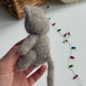 K&ouml;nnte beinhalten: Eine kleine, handgemachte Katzenfigur in Grau. Die Katze ist aus weichem, strukturiertem Garn gefertigt und hat einen langen Schwanz. Die Figur wird in einer Hand gehalten, mit einem verschwommenen Hintergrund aus einem Weidenkorb und Lichterketten.