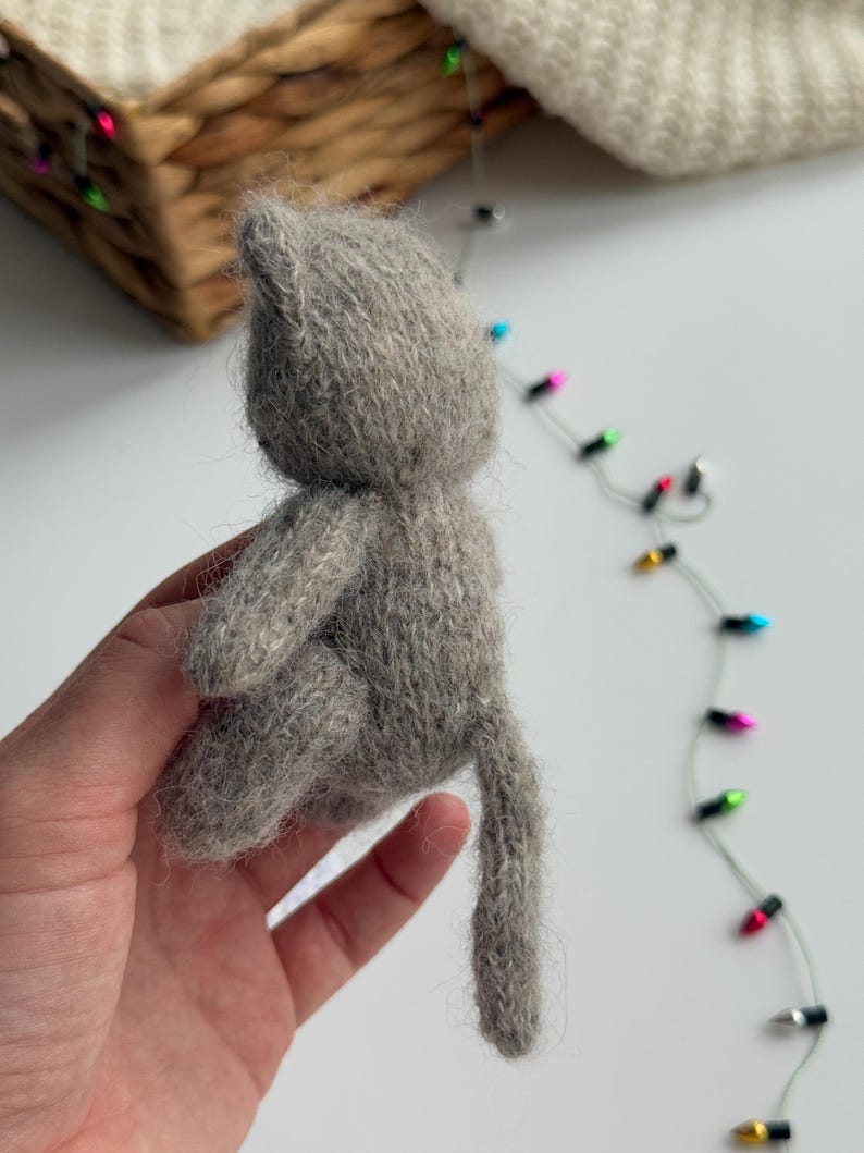 K&ouml;nnte beinhalten: Eine kleine, handgemachte Katzenfigur in Grau. Die Katze ist aus weichem, strukturiertem Garn gefertigt und hat einen langen Schwanz. Die Figur wird in einer Hand gehalten, mit einem verschwommenen Hintergrund aus einem Weidenkorb und Lichterketten.