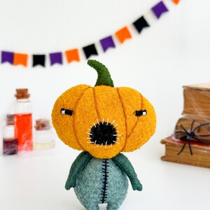 May include: A felt pumpkin character with a stitched mouth and eyes. The pumpkin is orange and the body is green. The character is standing on a white surface.