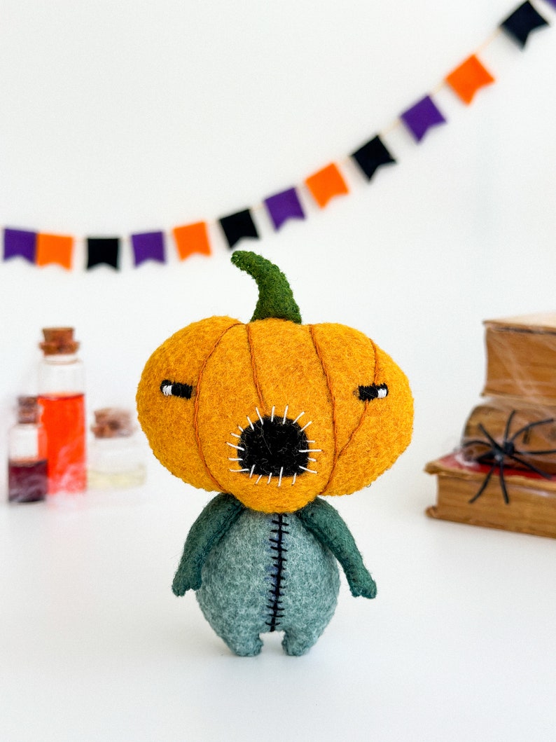 May include: A felt pumpkin character with a stitched mouth and eyes. The pumpkin is orange and the body is green. The character is standing on a white surface.