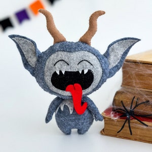 May include: A gray felt monster with horns, a big smile, and a red tongue sticking out. The monster is standing on a white surface next to a stack of old books with a black spider web on top.