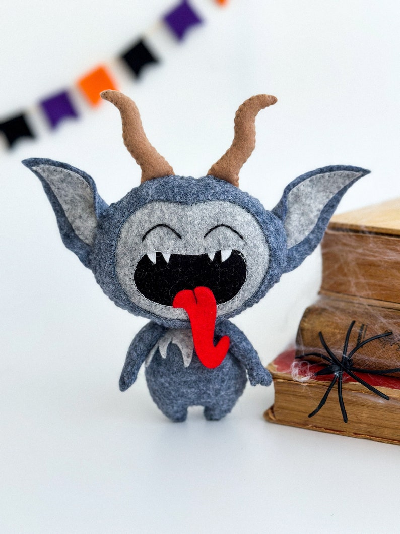 May include: A gray felt monster with horns, a big smile, and a red tongue sticking out. The monster is standing on a white surface next to a stack of old books with a black spider web on top.