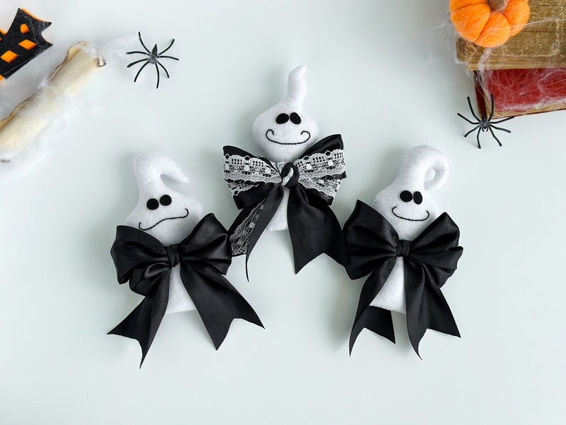 May include: Three white ghost ornaments with black bows. One ghost has a black and white lace bow. The ghosts are on a white background.
