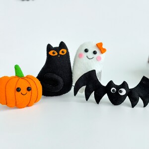 May include: A set of four felt Halloween decorations: a black cat, a white ghost with a pink bow, a black bat, and an orange pumpkin with a smiling face.