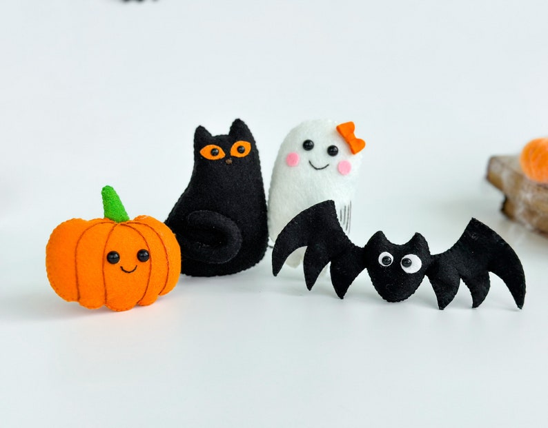 May include: A set of four felt Halloween decorations: a black cat, a white ghost with a pink bow, a black bat, and an orange pumpkin with a smiling face.