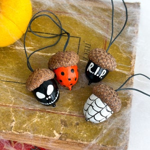K&ouml;nnte beinhalten: Vier Eichel-Ornamente, die mit Halloween-Motiven bemalt sind. Eine Eichel ist schwarz mit Totenkopf und Knochen gemalt, eine andere ist schwarz mit den Worten "RIP" bemalt, eine weitere ist orange und schwarz mit schwarzen Punkten bemalt und die letzte ist wei&szlig; mit einem Spinnennetz bemalt.