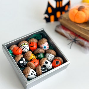 K&ouml;nnte beinhalten: Eine silberne Schachtel gef&uuml;llt mit bemalten Eicheln, die mit Halloween-Motiven verziert sind, darunter Geister, K&uuml;rbisse, Spinnen und ein Sch&auml;del. Die Eicheln sind in einem zuf&auml;lligen Muster angeordnet.