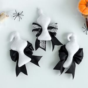 May include: Three white ghost-shaped ornaments with black bows. The bows are tied with black ribbon and white lace. The ornaments are on a white background.