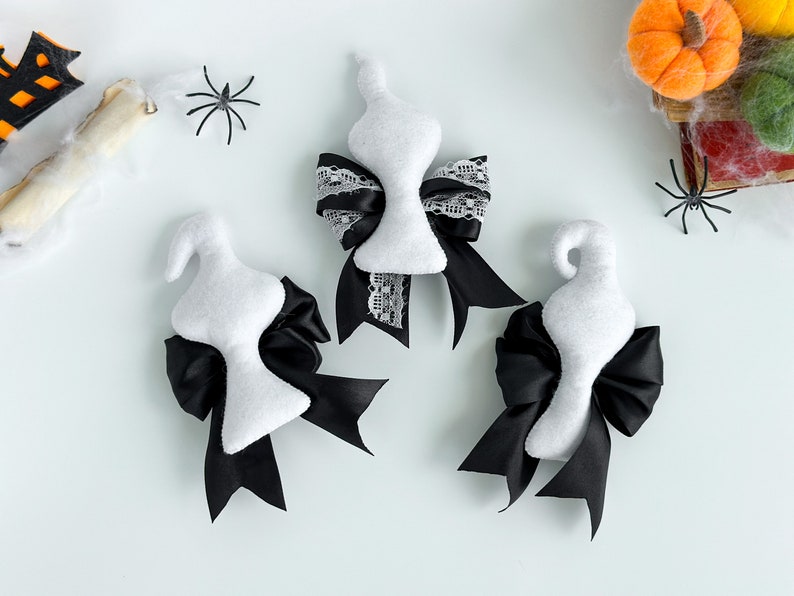 May include: Three white ghost-shaped ornaments with black bows. The bows are tied with black ribbon and white lace. The ornaments are on a white background.