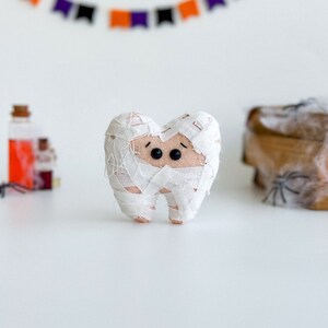 May include: A plush tooth wrapped in white bandages, resembling a mummy, with black button eyes. A Halloween-themed decoration. A banner with orange, purple, and black flags hangs in the background.