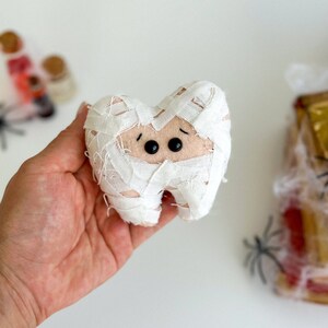 May include: A plush tooth wrapped in white fabric bandages, resembling a mummy. The tooth has black button eyes and a light tan face. The image suggests a Halloween or dental-themed craft.
