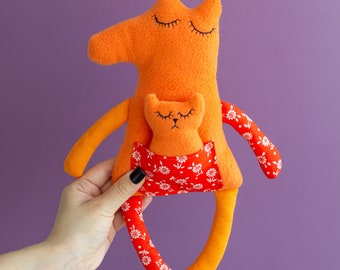 May include: A handmade orange plush toy with a smaller orange plush cat in its pocket. The toy has a red pocket with white floral print and orange and white striped legs.