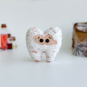 May include: A handmade Halloween-themed felt tooth, wrapped in white bandages to resemble a mummy. The tooth has black button eyes and a small, stitched mouth. The background includes Halloween decorations.