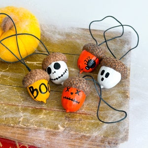 K&ouml;nnte beinhalten: F&uuml;nf Halloween-Ackerm&auml;nnchen, die mit verschiedenen Motiven bemalt sind. Die Eicheln sind mit schwarzem Garn aufgereiht und zeigen ein Gespenst, eine Spinne, einen Sch&auml;del, einen K&uuml;rbis und ein "BOO"-Design.