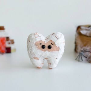 May include: A plush tooth dressed as a mummy for Halloween. The tooth is wrapped in white bandages, with black button eyes and a light brown face. A Halloween banner is in the background.