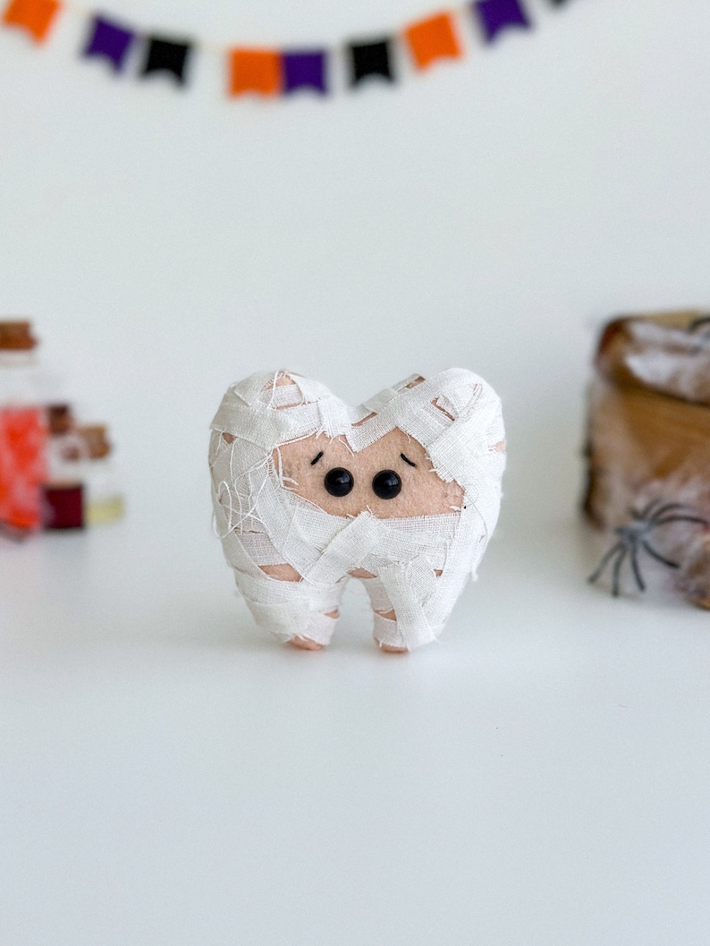 May include: A plush tooth dressed as a mummy for Halloween. The tooth is wrapped in white bandages, with black button eyes and a light brown face. A Halloween banner is in the background.
